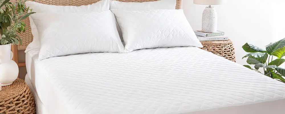A bed is dressed with a mattress protector and two pillows with pillow protectors.